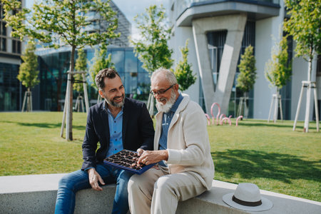 Senior dad and adult son sharing a box of sweets, chocolates on park bench.の写真素材