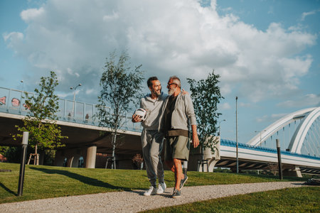 Sporty father and adult son walking home after football match in park.の写真素材