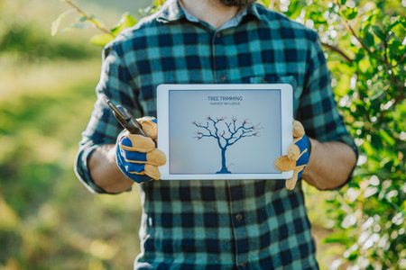 Farmer with tablet standing outdoors in orchard, learning how to trim a tree.