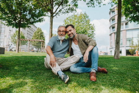 Father and adult son sitting on grass in park, sharing a story from the past.の写真素材