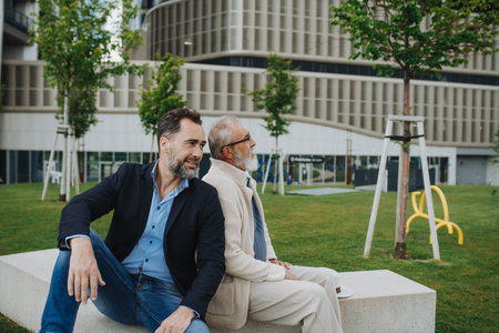 Father and adult son sitting on bench in park, sharing peaceful moment.の写真素材