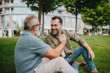 Father and adult son sitting on grass in park, sharing a story from the past.の写真素材