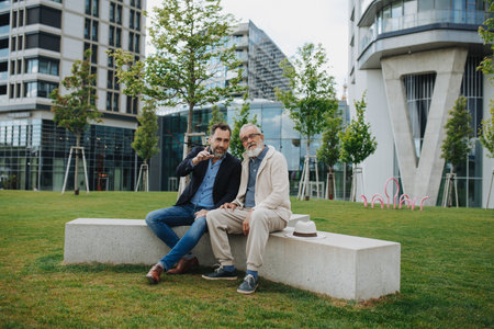 Father and adult son sitting on bench in park, sharing peaceful moment.の写真素材