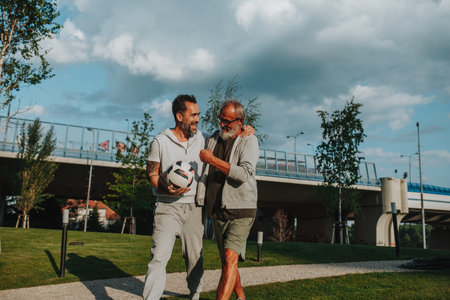 Sporty father and adult son walking home after football match in park.の写真素材