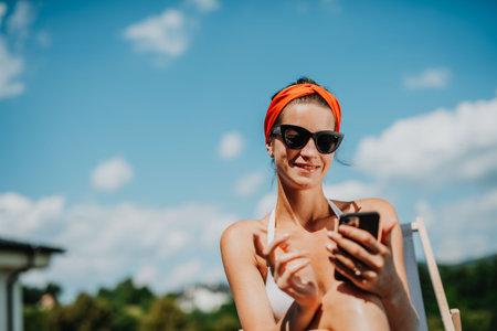 Young woman sitting by swimming pool, scrolling on smartphone.の写真素材