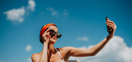 Young woman sitting by swimming pool outdoors in backyard garden, taking selfie.の写真素材