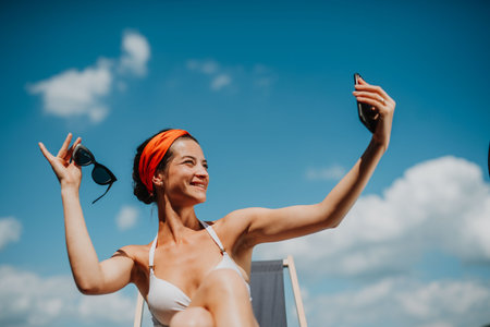 Young woman sitting by swimming pool outdoors in backyard garden, taking selfie.の写真素材