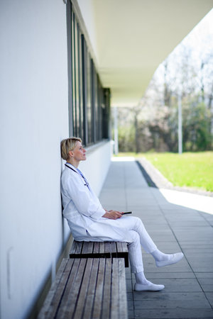 Young female doctor taking a break, sitting on bench in front of hospital.の写真素材
