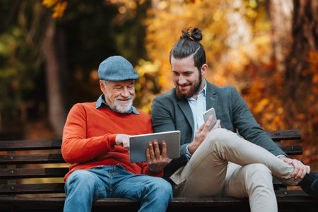 Senior father and his son sitting on bench by lake in nature, looking at tablet.の写真素材