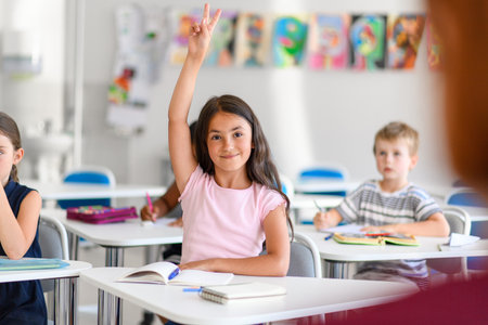 School girl sitting at the desk in classroom on the lesson, raising hands.