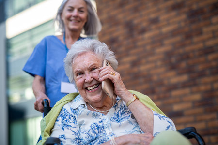 Older nurse pushing senior woman in wheelchair while making phone call.