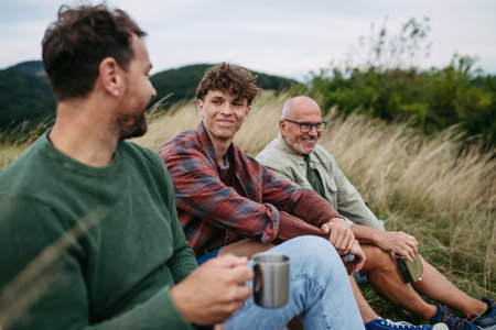 Grandfather, father and teen boy hiking togeter in nature, resting on meadow.