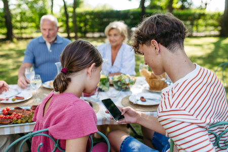 Siblings looking at phone during family barbecue in summer garden.の写真素材