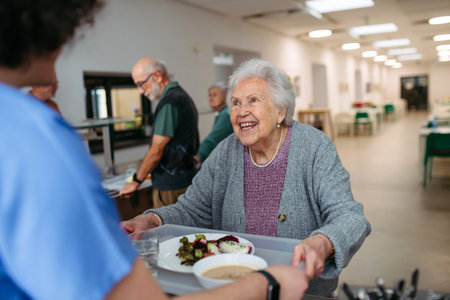 Senior woman having lunch with supportive caregiver in community center cafeteria.の写真素材
