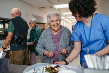 Senior woman having lunch with supportive caregiver in community center cafeteria.の写真素材