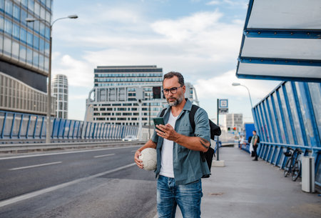 Bearded man with ball under arm using phone while waiting for bus.の写真素材