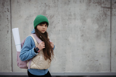 Outdoor portrait of sad young girl with beanie hat and backpack. Girl with long hair going to...