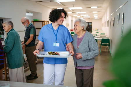 Senior woman having lunch with supportive caregiver in community center cafeteria.の写真素材