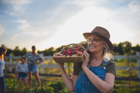 Older female farmer carrying basket with homegrown vegetables outdoors at community farm.の写真素材