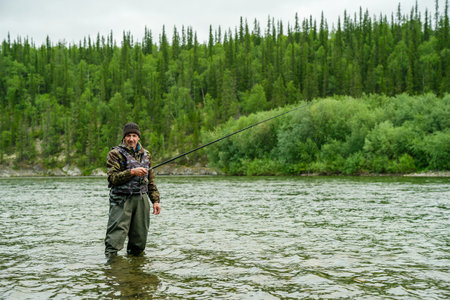 A fisherman stands in a river near a forest with a fishing rod
