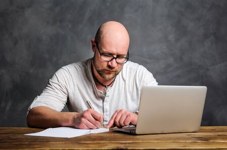 A man at a computer writes in a notebook. A man works with a laptop. A man is sitting at the table....