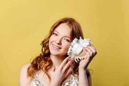 A young woman put a sea shell to her ear. A girl with red hair smiles and looks at the camera,...