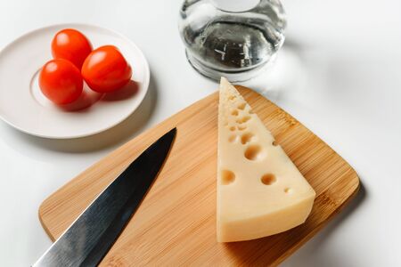 composition with a triangular piece of cheese, a cook knife and tomatoes on a white surface.の写真素材