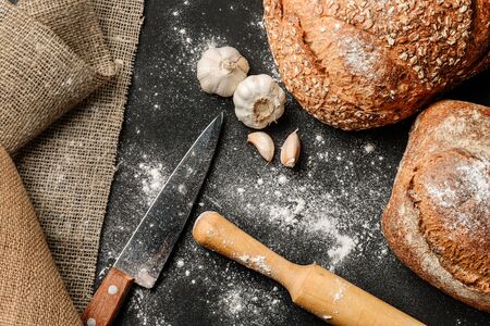 Flat lay composition with homemade bread, garlic, a kitchen knife, burlap and a rolling pin on a flour-covered table. The concept of a rural bakery.の写真素材