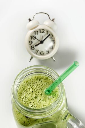 Green smoothie in a glass jar with a straw closeup with an alarm clock on a white background. Healthy smoothie breakfast. Healthy eating concept.の写真素材