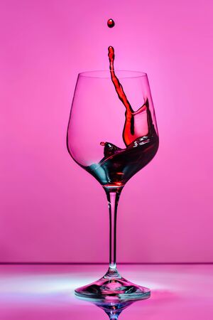 Red wine splashing in a glass on a pink background with blue reflexes and reflection in a glass countertop.の写真素材