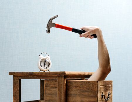 The alarm clock stands on the bedside table, a hand with a hammer, lifted above the alarm clock, is stuck out of an extended drawer. Early morning concept.の写真素材