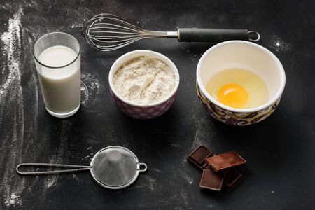 Ingredients for making muffins on a black surface sprinkled with flour. Flour, egg, whisk,...