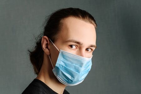 A young man in a medical mask looks at the camera, standing on a dark background.