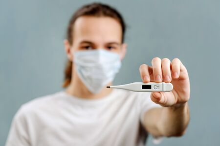 A young man in a medical mask holds a thermometer on his arm extended to the camera. Selective...