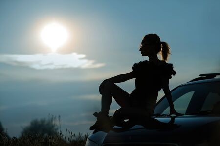 Silhouette of a girl in sunglasses sitting on the hood of a car against the sunset sky.の写真素材