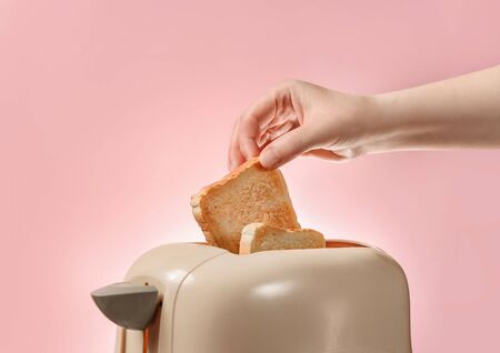 A female hand takes out toasted bread toast from a toaster. Toaster with ready-made toasts and a female hand isolated on a pink background with a copy space.の写真素材