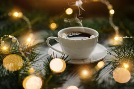 A cup of hot coffee with marshmallows among the fir branches and lights of festive garlands. Winter...