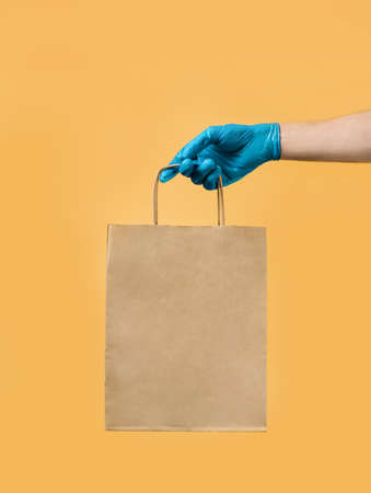 hand in a blue latex glove holds a craft bag on a yellow background. Safe delivery concept.