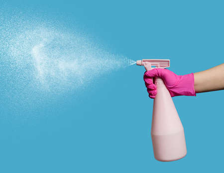 A hand in a latex glove holds a spray bottle on a blue background. Household chores. Minimalistic composition with a spray gun and a stream of water on a background with a copy space.の写真素材