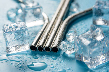 Metal cocktail pipes on a blue background among melting ice ubiks. Photo of a steel cocktail tube close up with selective focus.の写真素材