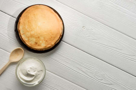 A plate of pancakes and a bowl of sour cream on a white wood background with copy space. Russian...