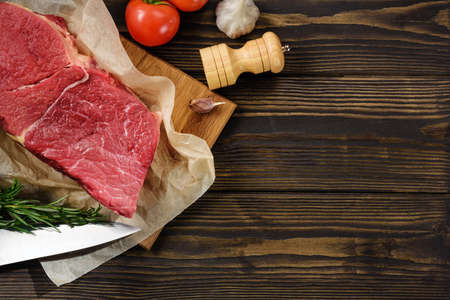 A piece of fresh beef, a tomato knife and seasoning on a wooden table with a copy space. Flat lay...