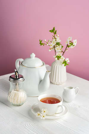 Spring composition with white tea utensils on a pink background. Cup with tea, teapot, milk jug and...