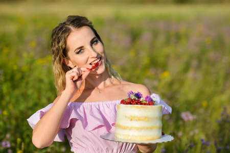 caucasian freckled woman holding a cake and eating berries posing outdoors on a sunny day.の写真素材
