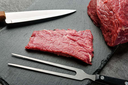 A piece of raw meat, a barbecue fork and a knife on a dark background close-up.の写真素材