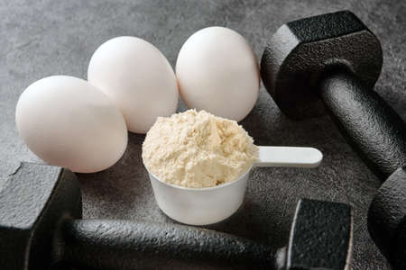 A scoop of egg white, eggs and a pair of black dumbbells on a dark background close-up.の写真素材
