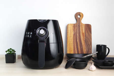 a black deep fryer or oil free fryer appliance, mug, dish and wooden tray are on the wooden table in the kitchen with a small plant in the pot ( air fryer ) with background of white cement wallの写真素材