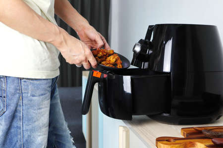 Right hand putting the fried chicken into the tray of the black deep fryer or oil free fryer appliance which is on the wooden table in the white kitchen ( air fryer )の写真素材