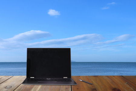 personal laptop notebook computer is put on the wooden table with a pen with background of beautiful sea and blue sky with clouds during working from home to avoid covid19 crisis on summer holidayの写真素材