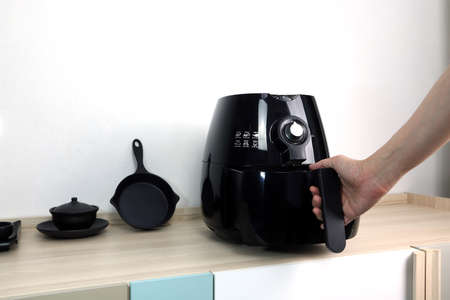 the right hand is holding the black air fryer or oil free fryer appliance which is on the wooden table in the white kitchen with white cement wall background for cooking the dinner for family membersの写真素材